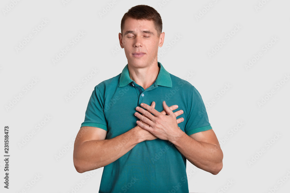 Acknowledgement and body language concept. Calm handsome man in bright t shirt, keeps hands on chest, expresses gratitude, being moved by heart piercing story, models against white background Acknowledgement and body language concept. Calm handsome man in bright t shirt, keeps hands on chest, expresses gratitude, being moved by heart piercing story, models against white background