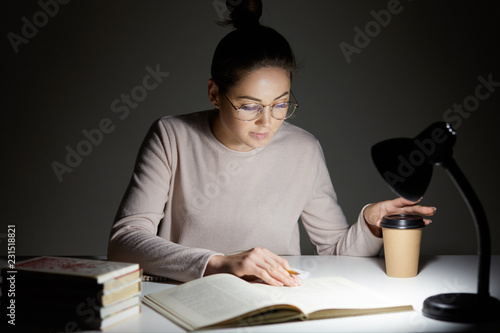 Isolated dark shot of concentrated female student prepares for upcoming exam, underlines information with pencil in textbook, uses lamp and optical round glasses for good vision, poses at desktop photo