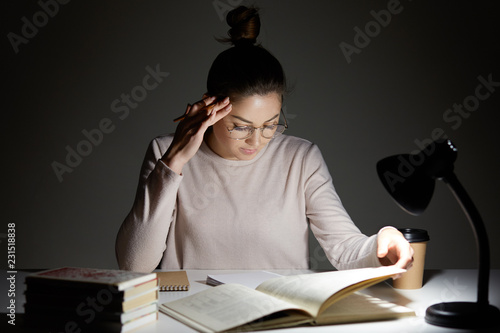 Horizontal shot of serious young European woman with hair bun, focused into book, learns material, writes down notes in notebook, sits at workplace with reading lamp and aromatic takeaway coffee photo