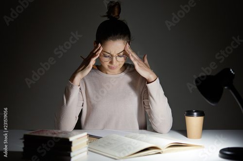 Tired stressful woman keeps hands on head, suffers from headache after working all night, tries to conecntrate, continue reading, dressed in casual clothes, wears transparent glasses enjoys cappuccino photo