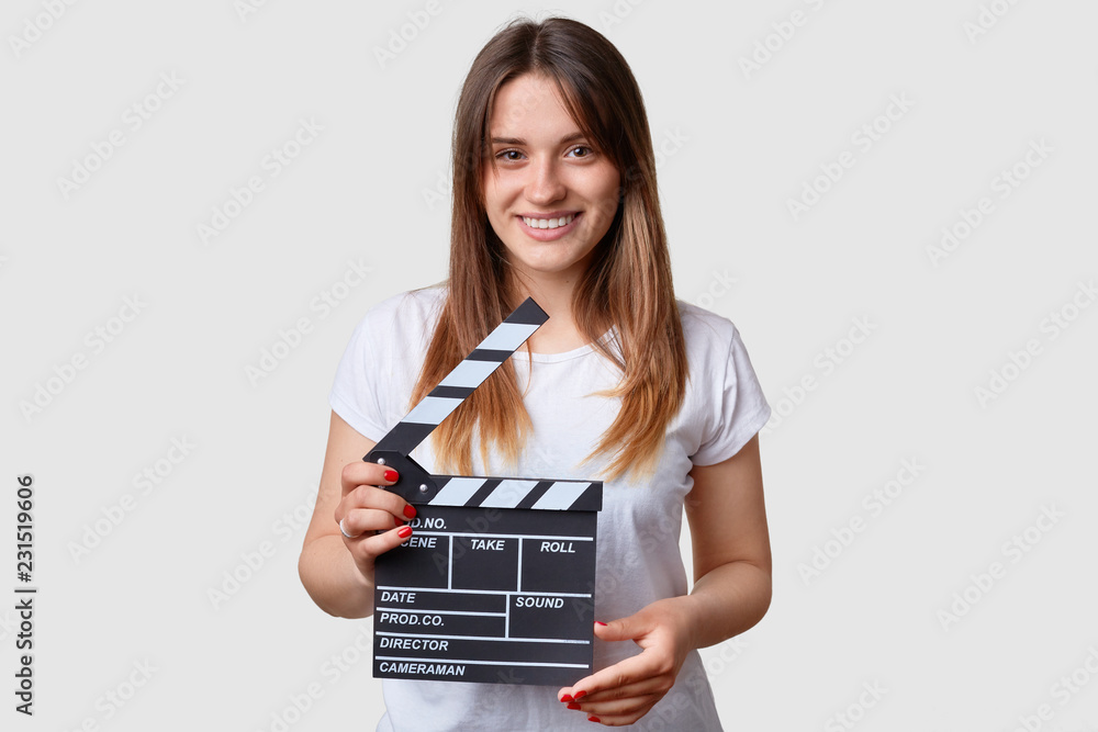 Film productivity concept. Beautiful pleased smiling young woman holds moving clapper board, prepares to shoot new scene, dressed in white casual t shirt, has practice as actress, models indoor.