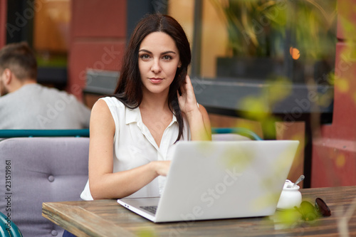 Outdoor shot of beautiful brunette woman in white blouse watches educational webinar to get more knowledge, sits in front of opened laptop in cafeteria, has e learning, connected to free internet