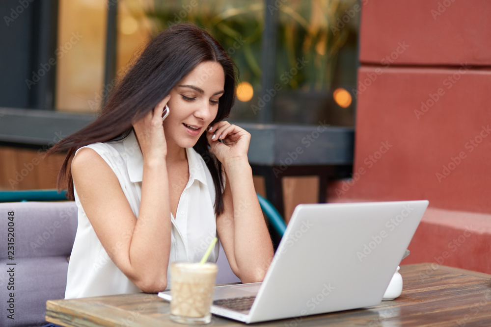 Fototapeta premium Ujęcie atrakcyjnej kobiety o ciemnych długich włosach, rozmawia z partnerem biznesowym przez sieć komórkową, używa laptopa do surfowania po Internecie i pracy na odległość, pije koktajl, pozuje w przytulnej kawiarni na świeżym powietrzu