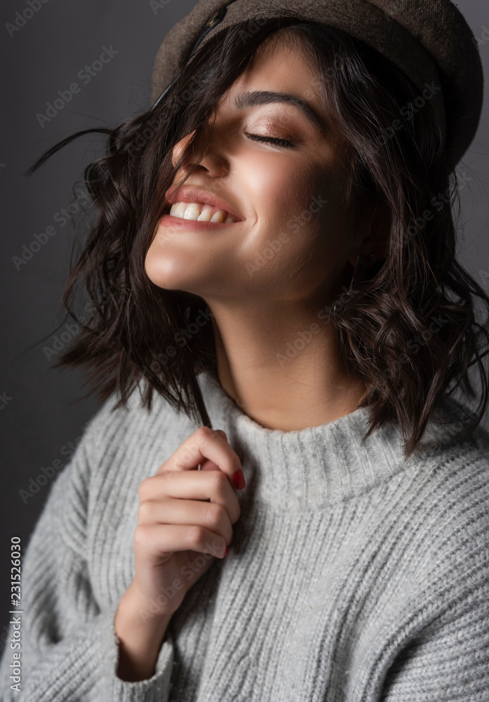 Beautiful smiling brunette girl wearing a casual style knitted sweater and cap touches her hair with her hand on gray background. Natural nude makeup. Fashion, advertising design. Close up. Beautiful smiling brunette girl wearing a casual style knitted sweater and cap touches her hair with her hand on gray background. Natural nude makeup. Fashion, advertising design. Close up.
