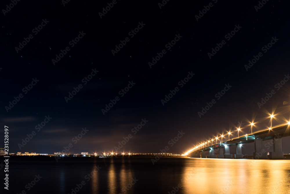Fototapeta premium Night cityscape with luminous bridge over wide river and starry dark sky
