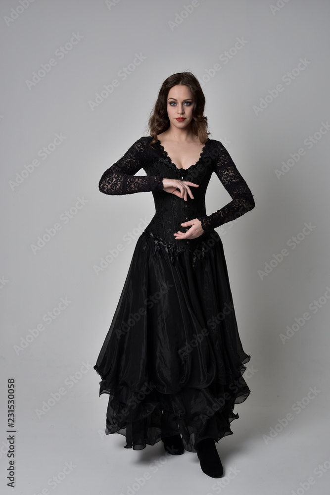 full length portrait of brunette girl wearing long black gown with corset. standing pose on grey studio background. full length portrait of brunette girl wearing long black gown with corset. standing pose on grey studio background.