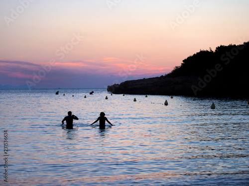 A sunset bay in menorca with purple and orange sky with an unidentifiable couple in silhouette in the dark blue calm evening sea