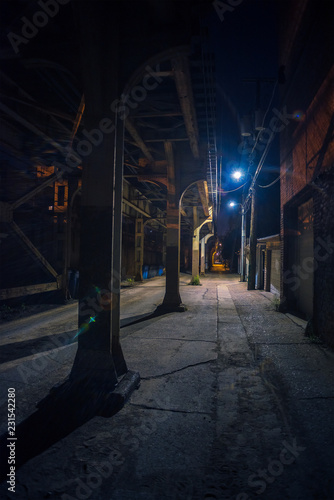 Dark and scary downtown urban city street alley under an eerie and illuminated vintage industrial railroad subway bridge at night