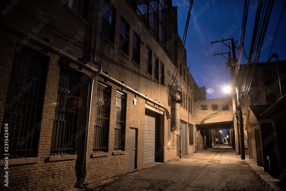 Dark and scary downtown urban city street alley scene with an eerie vintage industrial warehouse factory skyway at night