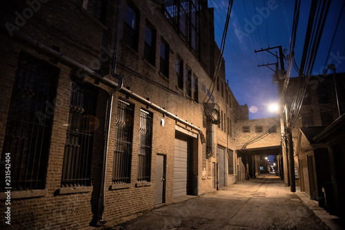 Dark and scary downtown urban city street alley scene with an eerie vintage industrial warehouse factory skyway at night