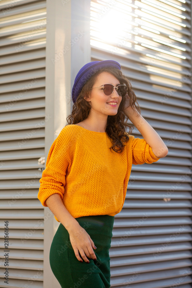 Street portrait of smiling model wearing blue hat and yellow knitted sweater posing in sun rays Street portrait of smiling model wearing blue hat and yellow knitted sweater posing in sun rays
