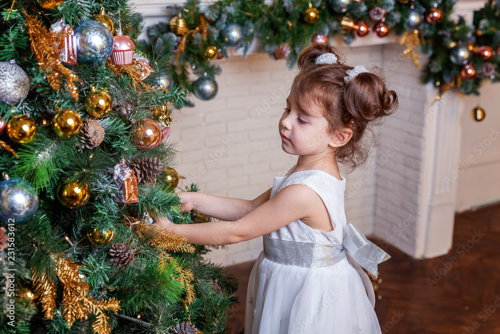 Little girl decorating Christmas tree on Christmas eve at home. Young kid in light bedroom with winter decoration. Happy family at home. Christmas New Year december time for celebration concept