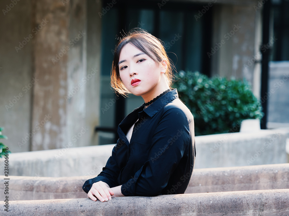 Fototapeta premium Beautiful Chinese young girl wearing black shirt looking and smiling at camera in summer city with back light and building background. Outdoor fashion portrait of glamour sexy stylish lady.