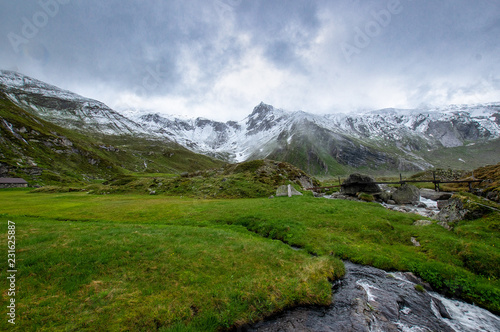 Schnee auf Berggipfeln der Alpen mit einen Fluß und grüner Wiese im Vordergrund