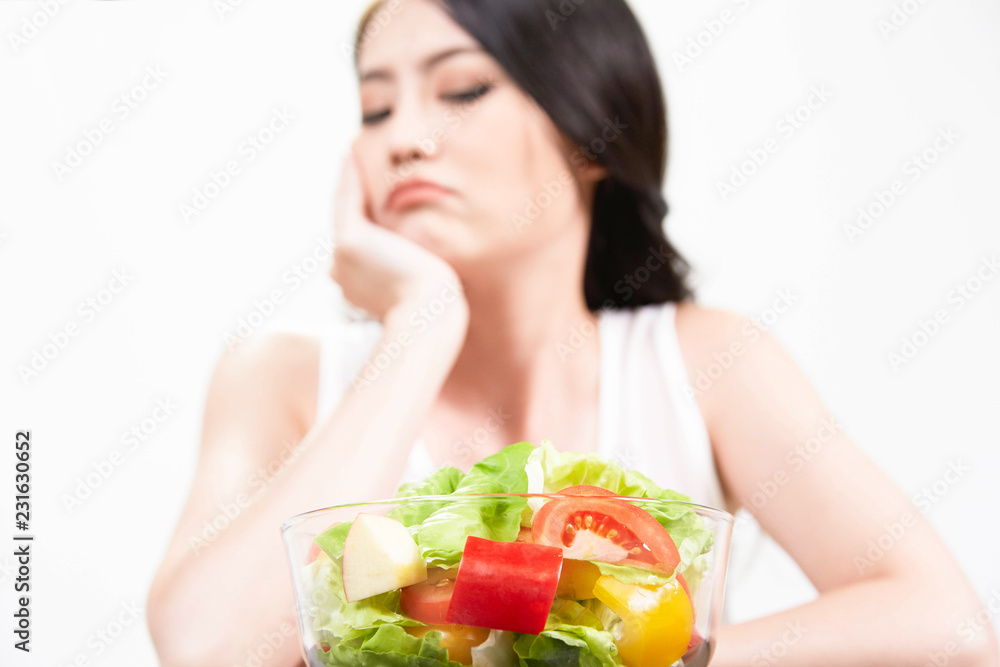 Unhappy  Asian women is on dieting time looking at green apple, red apple, kiwi, blueberries, tomato,bell peppers in transparent bowl her do not want to eat vegetables and dislike taste of salad...