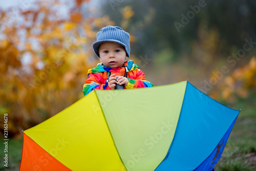 Sweet funny child with rainboy coat and multicolored umbrella jumping on puddles iand playing outdoors photo