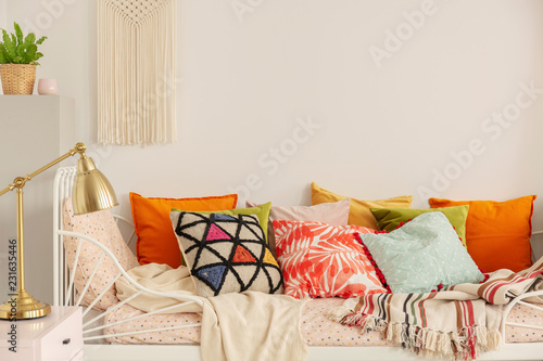 Patterned and colorful pillows on comfortable white bed in stylish bedroom interior with golden lamp on bedside table and handmade macrame on white wall, real photo with copy space