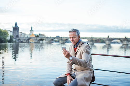 Sticker Mature businessman with smartphone sitting by river Vltava in Prague city