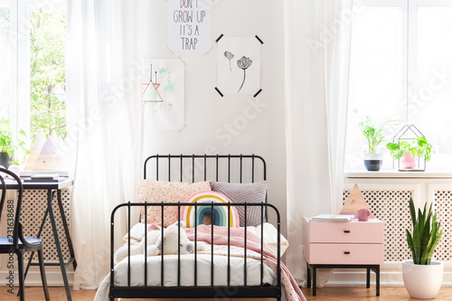 Plant and cabinet next to bed in white kid's room interior with posters and chair at desk. Real photo photo