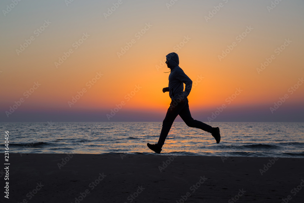 Alone man running on sand beach. Beautiful colorful sunset sky. Sportsman in tracksuit with hood. Enjoying sport in evening. Outdoor workout. Daily active lifestyle. Side view. Alone man running on sand beach. Beautiful colorful sunset sky. Sportsman in tracksuit with hood. Enjoying sport in evening. Outdoor workout. Daily active lifestyle. Side view.