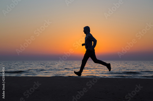 Alone man running on sand beach. Beautiful colorful sunset sky. Sportsman in tracksuit with hood. Enjoying sport in evening. Outdoor workout. Daily active lifestyle. Side view.