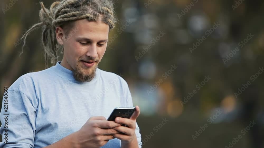 Extraordinary boy with dreads texting a message in the street, wearing casual shirt and gray t-shirt as using smartphone in lovely fall park Extraordinary boy with dreads texting a message in the street, wearing casual shirt and gray t-shirt as using smartphone in lovely fall park