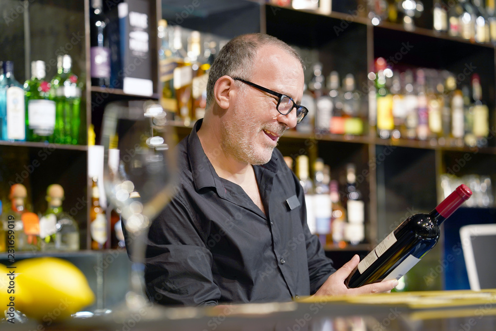 The barman is holding a bottle of red wine. The barman examines a bottle of red wine.The bartender pours red wine to the client in the hotel bar. The concept of service. Concentrate on the bartender. The barman is holding a bottle of red wine. The barman examines a bottle of red wine.The bartender pours red wine to the client in the hotel bar. The concept of service. Concentrate on the bartender.