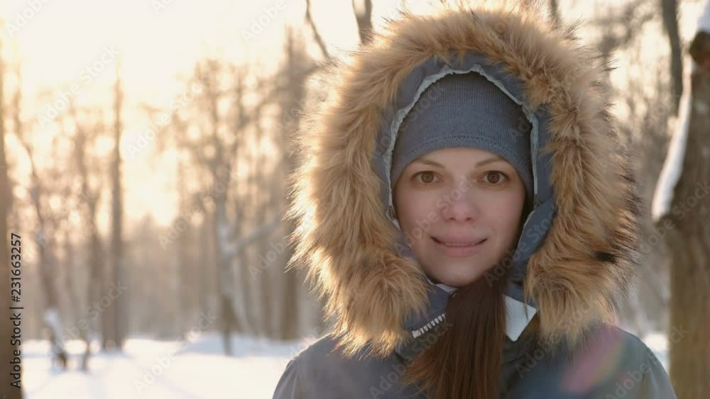 Close-up woman face in fur hood looking in camera and smiling in winter sunny park. Right on video Close-up woman face in fur hood looking in camera and smiling in winter sunny park. Right on video
