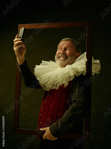 Official portrait of historical governor man with mobile phone from the golden age with corrugated round collar. Studio shot against dark wall. He making selfie photo photo