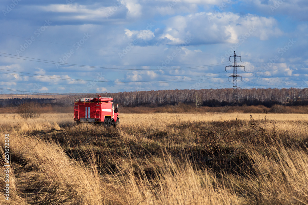 Fototapeta premium Red firetruck car Ural rides through the autumn field with yellow and faded grass against the blue sky and clouds. The concept of extinguishing forest fires in USA