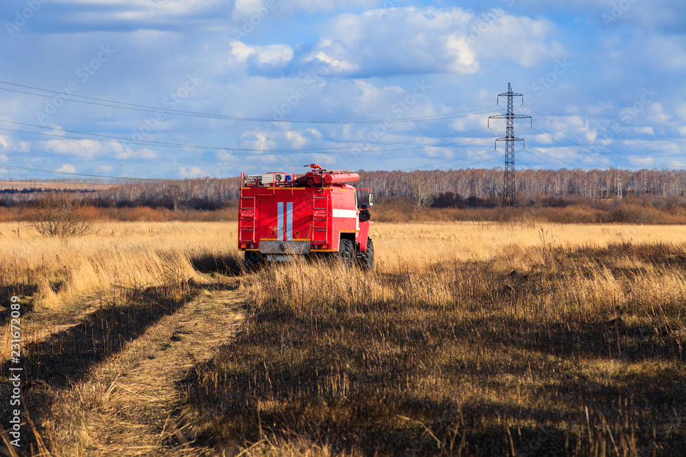 Fototapeta premium Red firetruck car Ural rides through the autumn field with yellow and faded grass against the blue sky and clouds. The concept of extinguishing forest fires in USA