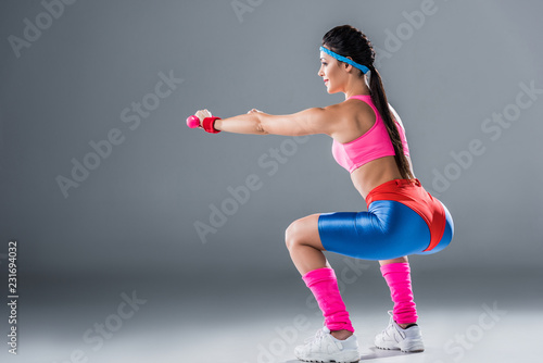 side view of sporty girl doing squats with dumbbells on grey