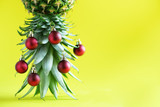 Creative Christmas tree made of pineapple and red bauble on yellow background, copy space. Greeting card, decoration for new year party. Holiday concept.