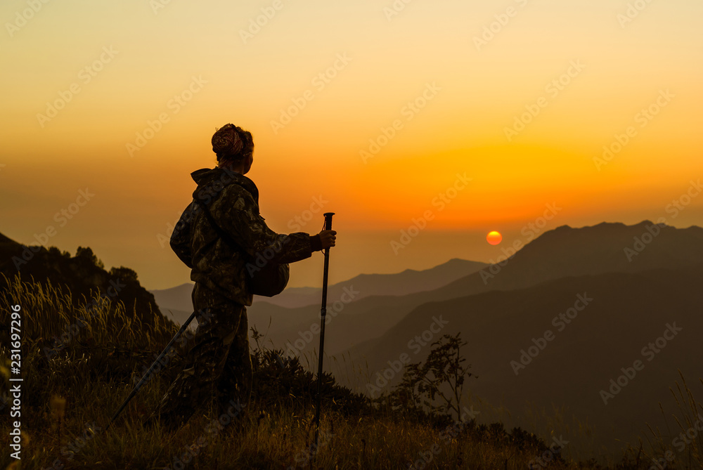 girl traveler with a camera case and trekking poles walks through the mountain pass and looks at the setting sun