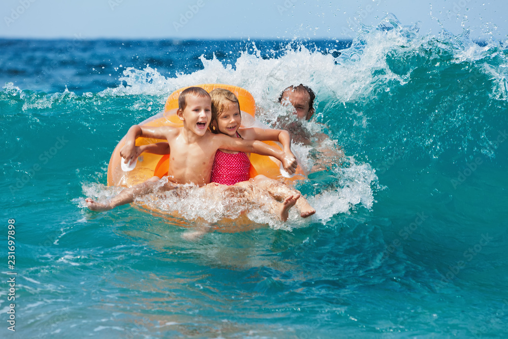 Happy kids have fun in sea surf on beach. Joyful couple of children on inflatable ring ride on breaking wave. Travel lifestyle, swimming activities in family summer camp. Vacations on tropical island 