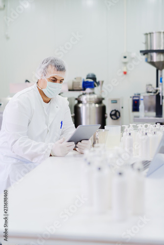 Picture of lotion bottles on production line. Bottles of cosmetic products in factory production line. Blurred picture of man using control panel in background.