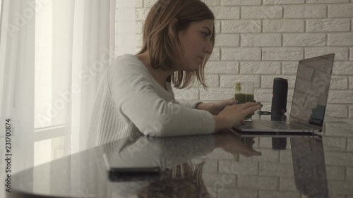 Woman working on her laptop at home sitting at the table drinking healthy green smoothie, then reaching and checking her phone photo