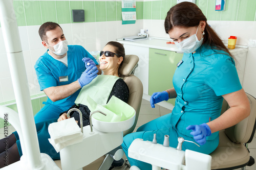 people  medicine  stomatology and health care concept - male dentist and assistant with dental curing light and mirror treating woman patient teeth at the real dental clinic office