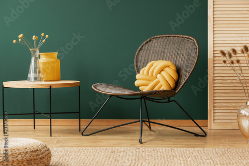 Glass vase with flowers on wooden coffee table next to yellow knot pillow on stylish wicker armchair, real photo with copy space on empty green wall