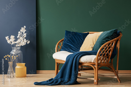 Glass vases with flowers next to comfortable rattan settee with blue, emerald and beige pillows and blanket, real photo with copy space on empty green wall