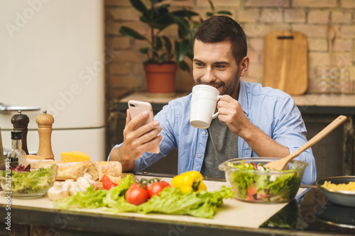 Man preparing delicious and healthy food in the home kitchen on a sunny day with coffee. Using phone. Thumbs up.