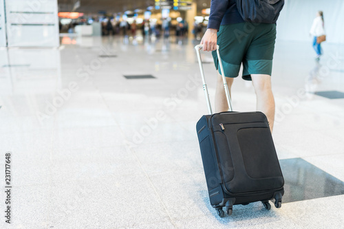 Young man wearing shorts and sneakers is walking through the airport holding a small-sized black suitcase with 4 wheels