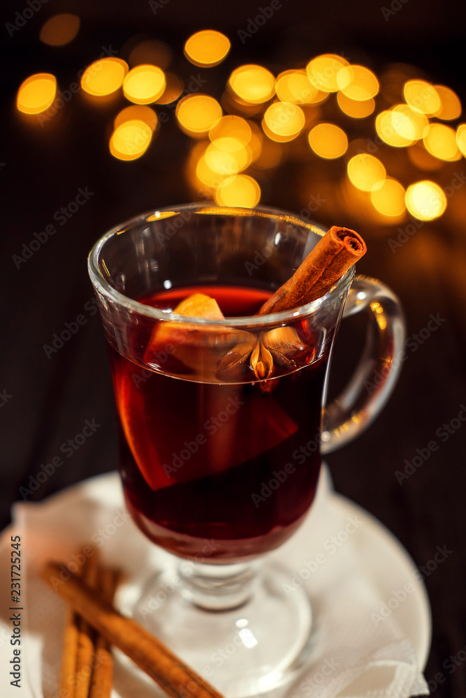 Fototapeta premium closeup glass of mulled wine with orange and cinnamon on dark black background, on white plate, Christmas lights