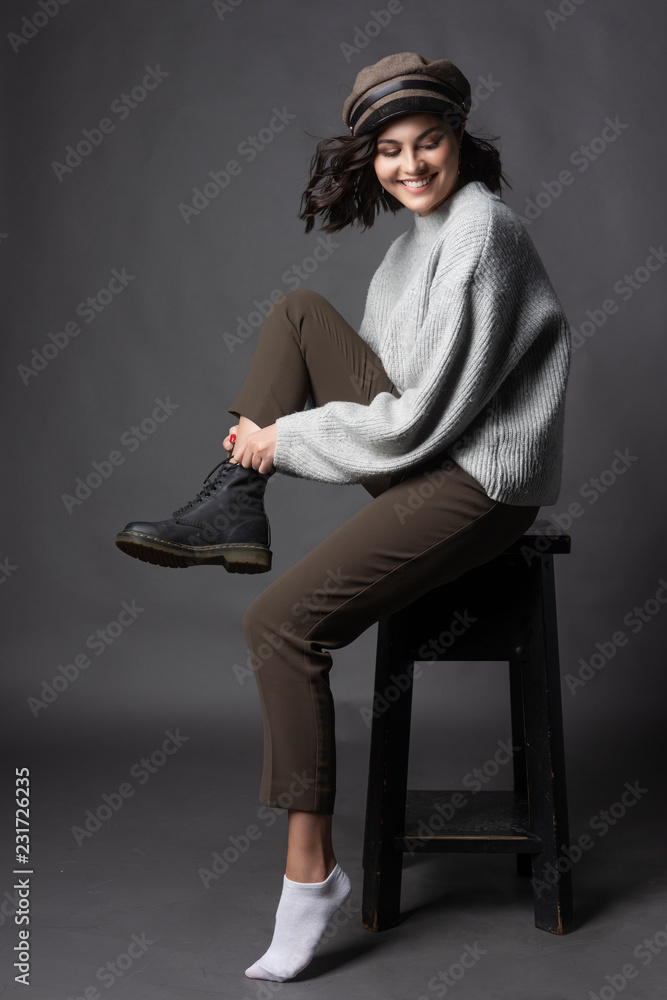 Beautiful smiling brunette girl wearing a casual style sweater, pants and cap puts a shoe on her leg on a gray background. Natural nude makeup. Fashion, advertising and commercial design Beautiful smiling brunette girl wearing a casual style sweater, pants and cap puts a shoe on her leg on a gray background. Natural nude makeup. Fashion, advertising and commercial design