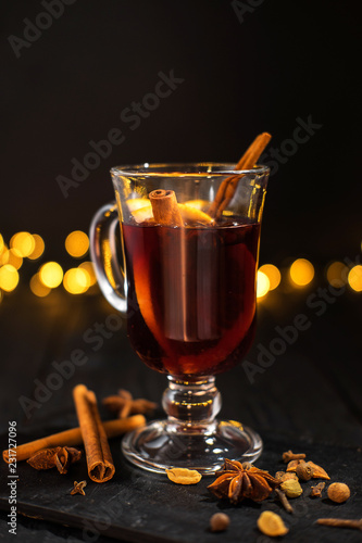 closeup glass of mulled wine with orange and cinnamon on dark black background, christmas lights, large yellow bokeh, mulled wine set