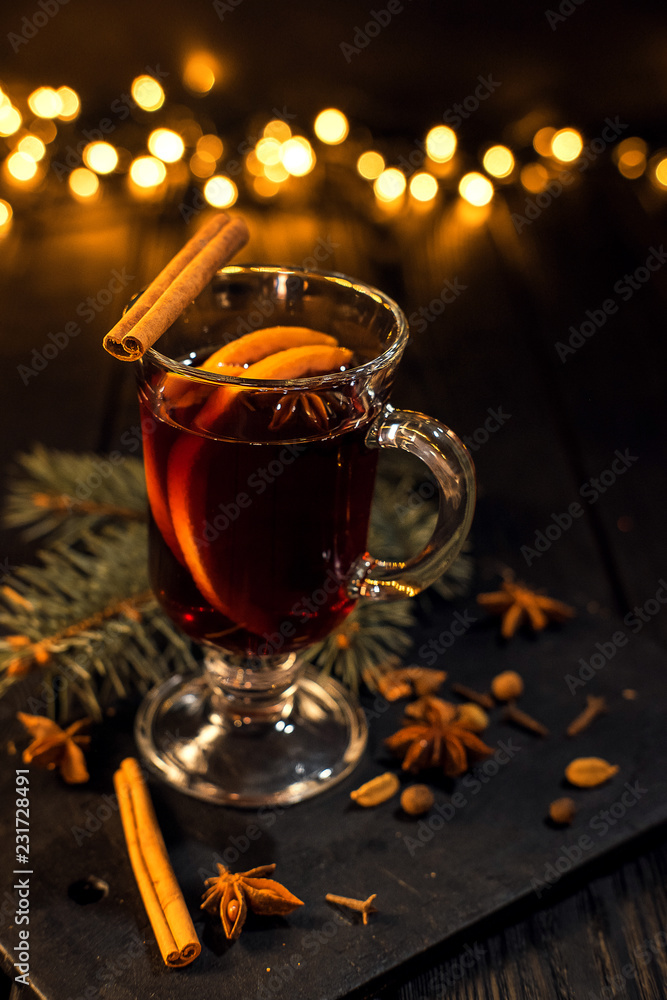 cinnamon lies in a glass, closeup glass of mulled wine with orange and cinnamon on dark black background, Christmas tree and lights, large yellow bokeh, mulled wine set cinnamon lies in a glass, closeup glass of mulled wine with orange and cinnamon on dark black background, Christmas tree and lights, large yellow bokeh, mulled wine set
