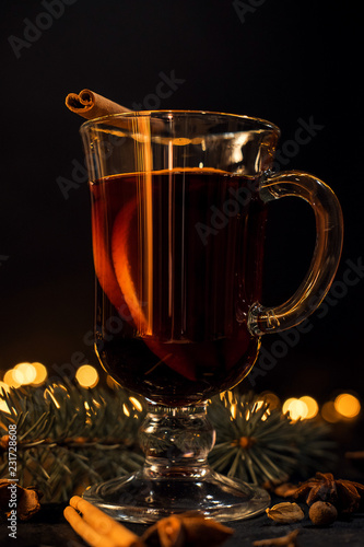 cinnamon lies in a glass, closeup glass of mulled wine with orange and cinnamon on dark black background, Christmas tree and lights, large yellow bokeh, mulled wine set
