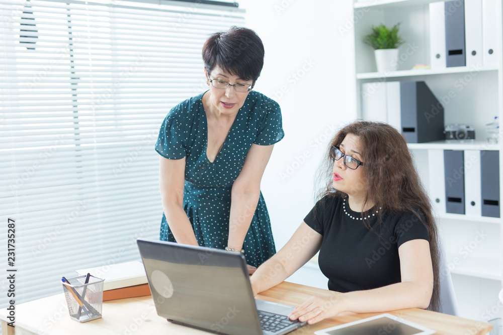 Business people, technology and communication concept - Mid-aged lady helping her female colleague with a laptop Business people, technology and communication concept - Mid-aged lady helping her female colleague with a laptop