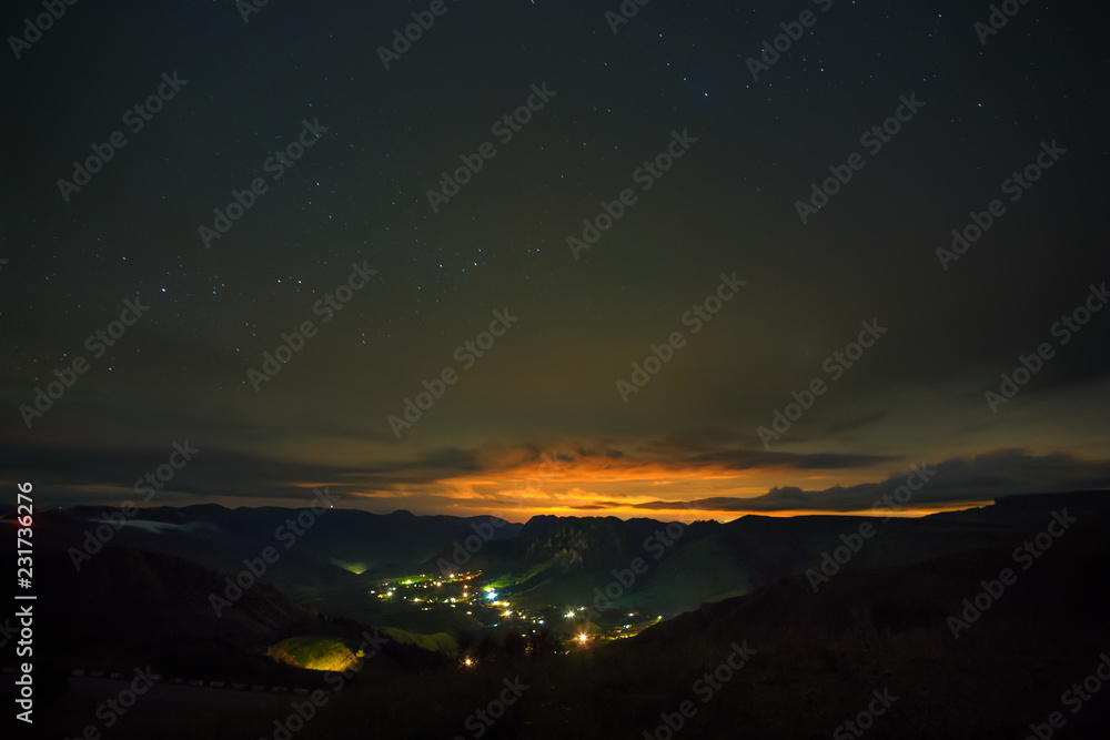 Bright stars shine through the clouds in the night sky over a city in a mountain valley. Overcast night in the summer.