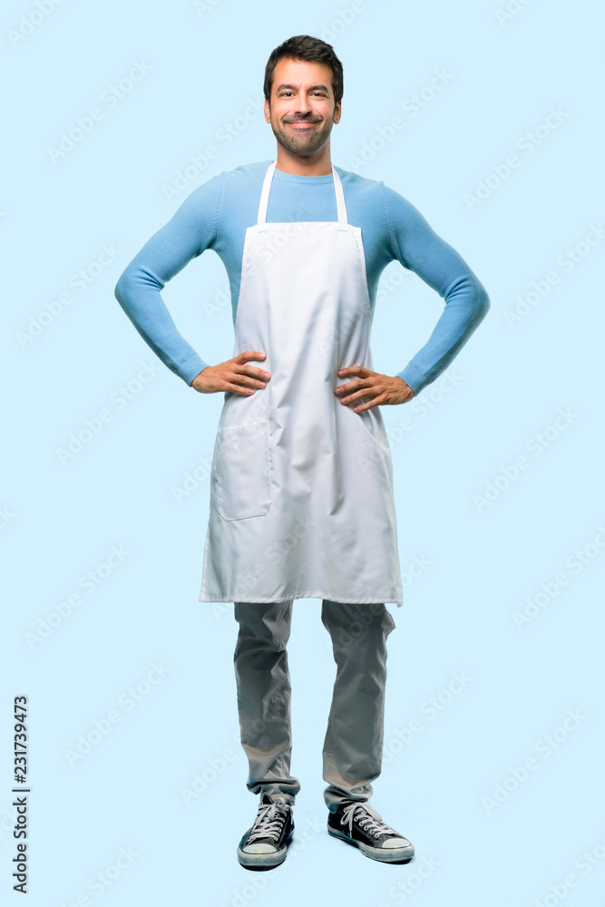 Full body of Man wearing an apron posing with arms at hip and laughing looking to the front on blue background